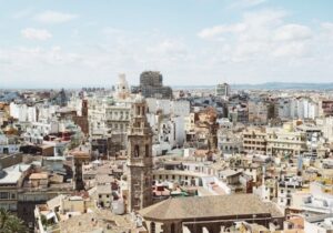 A cityscape view of Valencia, Spain, showing historic buildings, church towers, and modern structures under a partly cloudy sky.