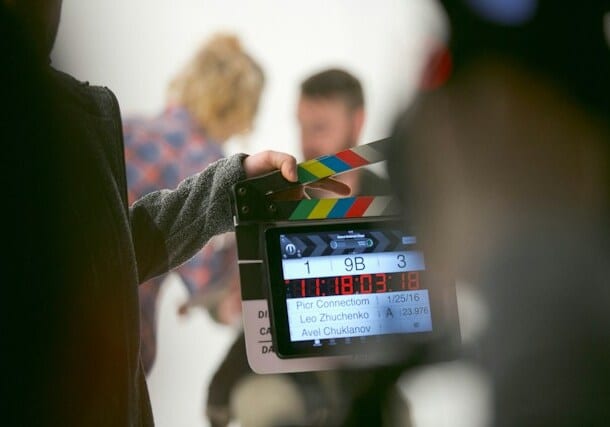 Film crew member holding a clapperboard on a set, with two blurred actors in the background.