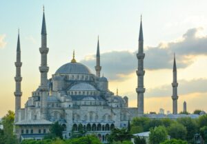 The Blue Mosque (Sultan Ahmed Mosque) in Istanbul at sunset, with its iconic domes and six towering minarets silhouetted against a pastel sky.