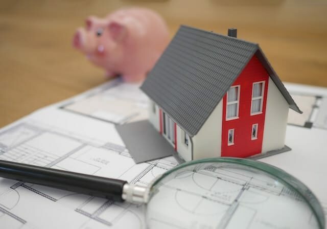 House plans, a magnifying glass and model house next to a piggy bank on a desk