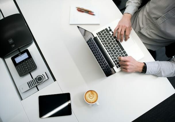 Top view of a person working on a laptop at a desk with a coffee, tablet, and notepad.