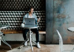 Man sitting at a table in a cafe with headphones on, working on a laptop - examples of country of residence