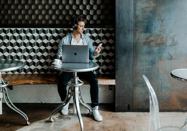 Man with headphones on sitting at a desk in a cafe working on his laptop and smartphone - Travel insurance for digital nomads and remote workers
