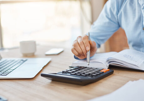Man in light blue shirt sitting at a desk working at a laptop and using a calculator - How to calculate crypto tax gains and losses