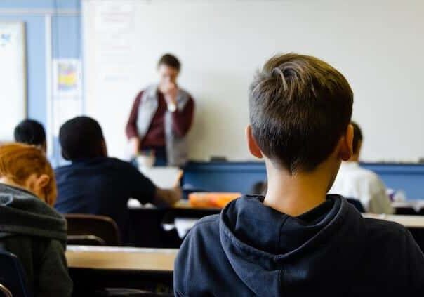 Students sitting in a classroom, facing a teacher who is speaking in front of a whiteboard.