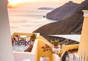 Sunset view over the sea from a charming terrace in Santorini, Greece, with yellow walls, outdoor seating, and a backdrop of cliffs.