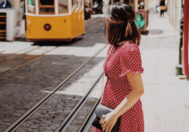 Woman in a red polka dot dress standing near tram tracks, looking at a yellow tram approaching.