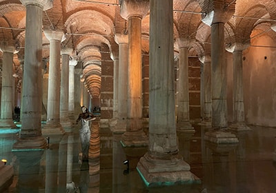 Interior of the Basilica Cistern in Istanbul, featuring rows of ancient marble columns reflected in shallow water, with ambient lighting creating a dramatic effect.