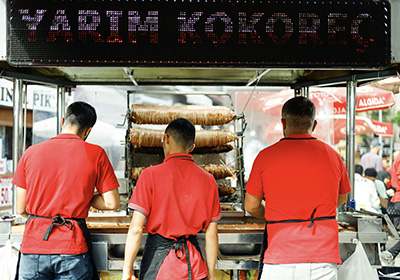 Street food vendors in red uniforms preparing kokoreç, a traditional Turkish grilled dish, at a busy outdoor food stall in Istanbul.