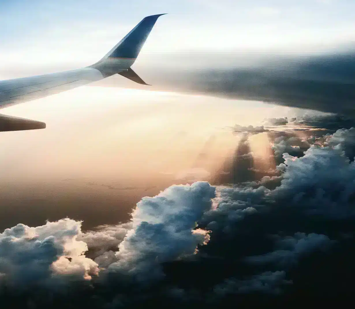 An airplane wing soaring above a blanket of fluffy white clouds against a clear blue sky.