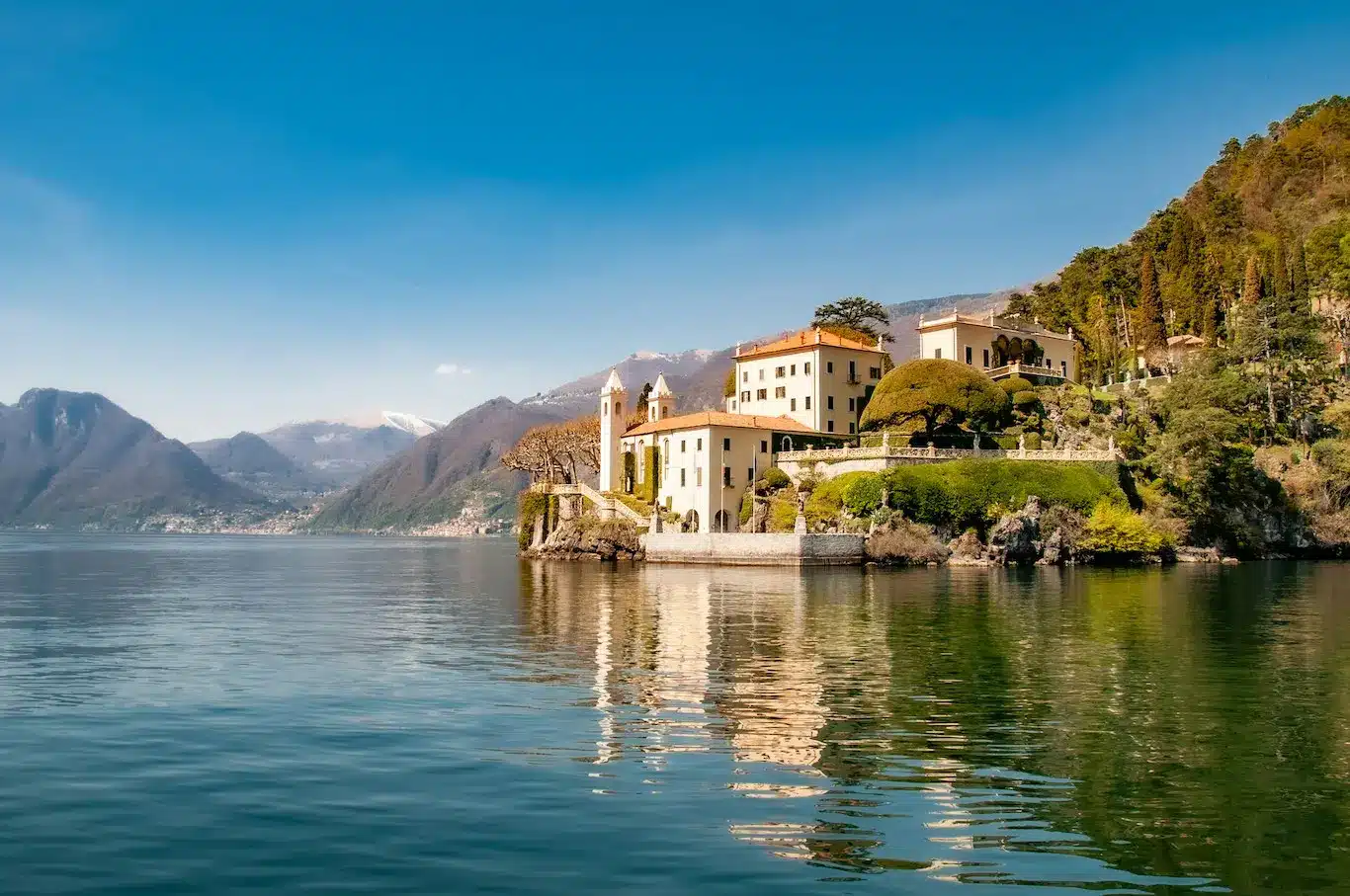 A serene lake reflecting a majestic castle surrounded by lush greenery under a clear blue sky.